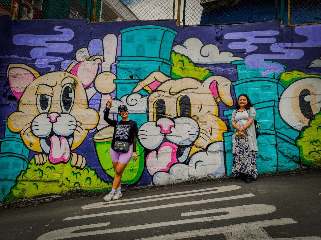 Dos personas posan frente a un mural urbano con ilustraciones de gatos coloridos